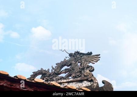 Una tradizionale scultura di drago, dettagliatamente dettagliata, si trova in cima a un'antica casa vietnamita, a simboleggiare potere e prosperità sotto un blu chiaro Foto Stock