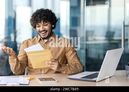 Un giovane eccitato in un abbigliamento casual da lavoro reagisce con gioia all'apertura di un pacchetto alla sua scrivania. Foto Stock