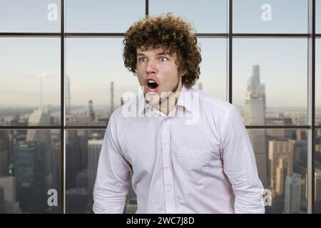 Giovane uomo caucasico con i capelli ricci che urla ad alta voce. Finestra a scacchi con vista sulla città sullo sfondo. Foto Stock