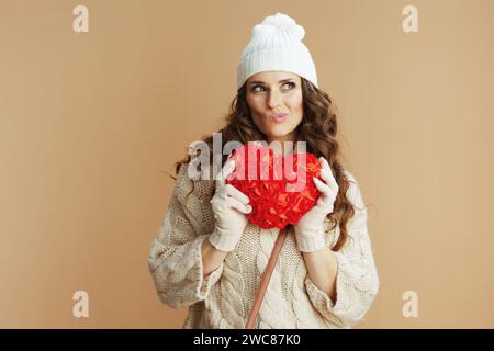 Ciao inverno. penosa donna di mezza età alla moda con maglione beige, guanti e cappello su sfondo beige con cuore rosso. Foto Stock