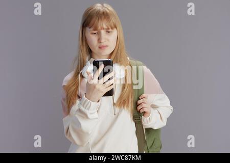infelice adolescente moderna in tuta beige con zaino, cuffie e smartphone isolato sul grigio. Foto Stock