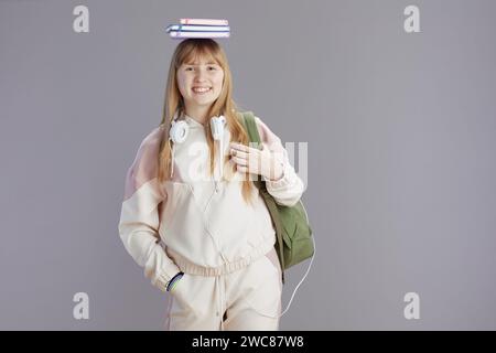 Ritratto di un'adolescente di tendenza sorridente in tuta beige con zaino, libri di lavoro e cuffie contro il grigio. Foto Stock