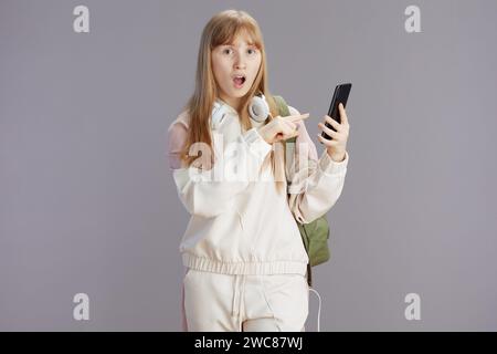una ragazza adolescente moderna sorprende con una tuta beige con zaino e cuffie che utilizza uno smartphone isolato su grigio. Foto Stock