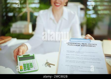 Un'attività immobiliare sostenibile. Primo piano su un elegante abitante femminile nel moderno ufficio verde in camicetta bianca con calcolatrice, appunti, documento e ke Foto Stock