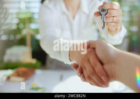 Un'attività immobiliare sostenibile. Primo piano su una giovane donna d'affari in un moderno ufficio verde in una blusa bianca con chiavi e cliente che stringe la mano. Foto Stock