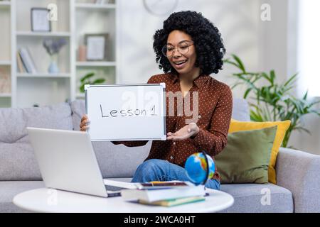 Una giovane insegnante afroamericana insegna online tramite videochiamata su laptop, tiene una lavagna con una lezione scritta in mano, sorride. Foto Stock