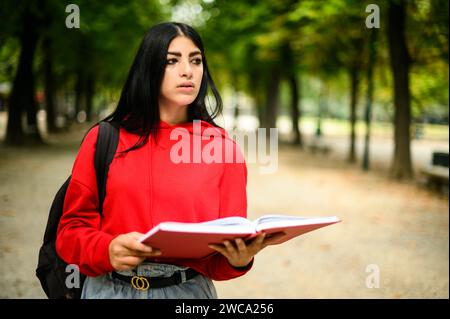 Una giovane donna con cappuccio rossa legge un libro mentre cammina in un parco lussureggiante Foto Stock