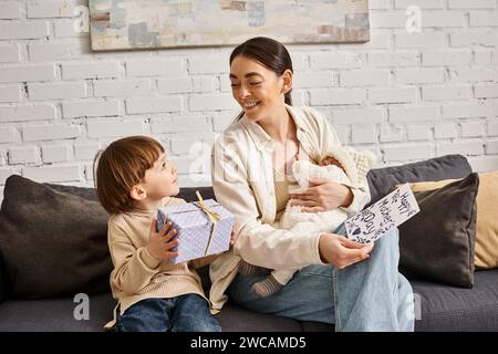 Donna attraente seduta sul divano con i figli con un regalo e un biglietto d'auguri per la festa della mamma Foto Stock