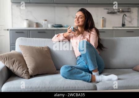 Donna contenta che assapora il caffè su un comodo divano Foto Stock