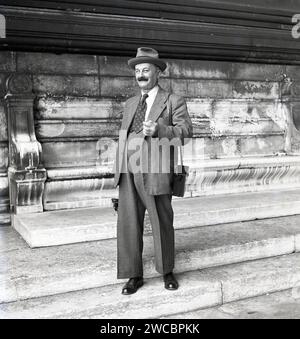 1950, storico, turista, gentiluomo con tuta e cravatta e cappello, con una macchina fotografica pieghevole e custodia in piedi su un gradino di pietra nella piazza della città, in Germania. Foto Stock