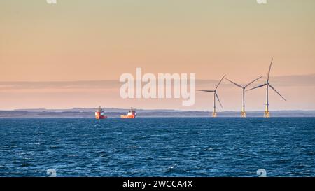Parco eolico nel mare vicino alla costa, con barche di rifornimento vicino, durante il tramonto con montagne e cielo limpido e colorato sullo sfondo. Foto Stock