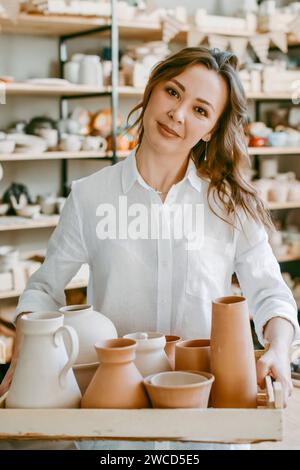 Una donna vasaio tiene una scatola con prodotti di argilla cruda. Una donna artigiana dimostra felicemente i suoi oggetti d'artigianato in un laboratorio di ceramica Foto Stock