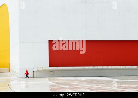 Museo del moderno Niemeyer Center con una donna in una giacca rossa che cammina di fronte ad esso, ad Aviles, Asturias, Spagna. Foto Stock