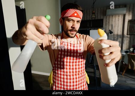 Giovane con grembiule e fascia che tiene due bottiglie di detersivi mentre si trova davanti alla fotocamera durante le faccende domestiche Foto Stock