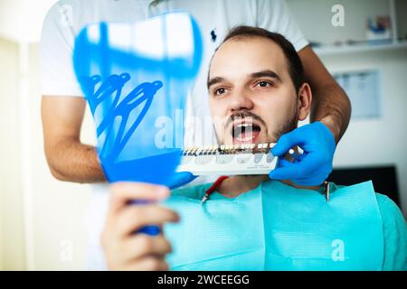 Il paziente al dentista guarda i suoi denti attraverso lo specchio, seleziona il colore dei denti dalla tavolozza dei denti Foto Stock