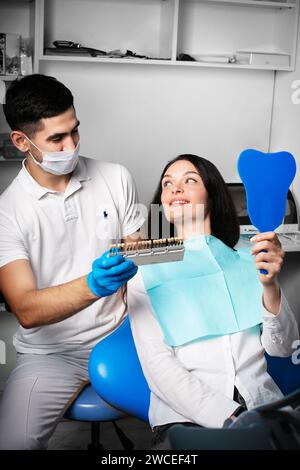 Una donna alla poltrona dentista guarda i suoi denti seleziona il colore dalla tavolozza dei denti Foto Stock