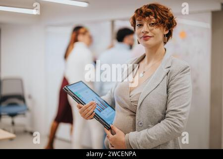 Splendida donna d'affari incinta rossa che si posa in ufficio mentre tiene il laptop. La sua squadra sta facendo il brainstorming alle spalle. Copia spazio. Foto Stock