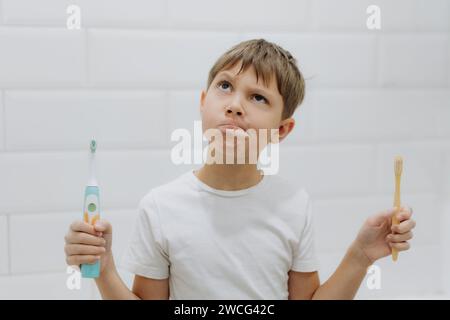 carino bambino di 8 anni che tiene in mano bambù e spazzolino elettrico per fare la scelta Foto Stock
