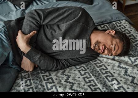 uomo barbuto che soffre mentre è sdraiato sul letto e soffre di mal di stomaco, dolore addominale Foto Stock