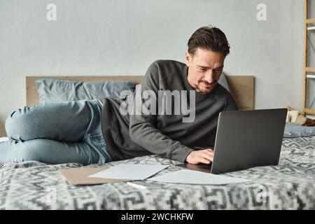 uomo concentrato e barbuto che utilizza un notebook mentre lavora in remoto da casa in camera da letto e documenti Foto Stock