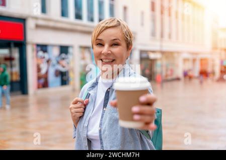 una donna felice in camicia blu con zaino e caffè sotto una passeggiata per una città moderna Foto Stock