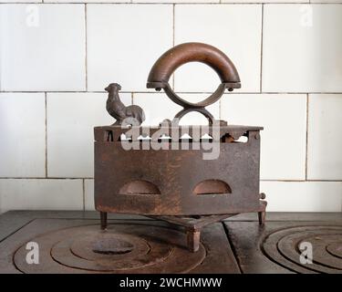 Ferro da stiro per abiti antichi, ferro da stiro per abiti del XIX secolo con una figura di gallo in cima, seduto sopra il forno a legna Foto Stock