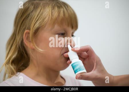 la madre spruzza il naso contro la febbre di sua figlia, la bambina viene trattata a casa ed è in pigiama - sfondo sfocato. Foto Stock