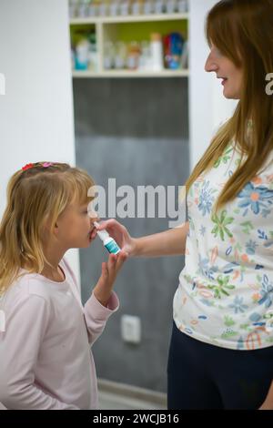 la madre spruzza il naso contro la febbre di sua figlia, la bambina viene trattata a casa ed è in pigiama - sfondo sfocato. Foto Stock