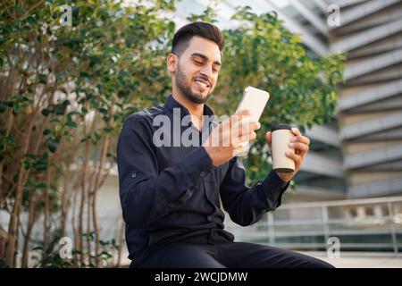 uomo d'affari arabo che parla con lo smartphone, videochiama bevendo un caffè all'aperto Foto Stock