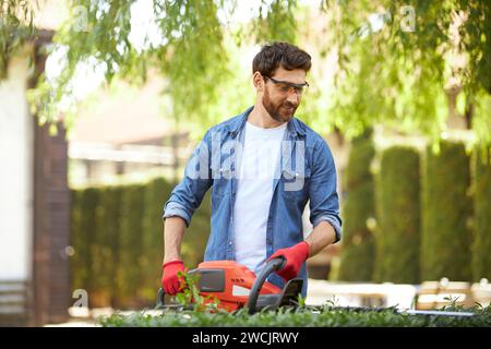 Il giardiniere maschio sorridente brunet taglia il cespuglio troppo coltivato con un tagliasiepi nelle giornate di sole. Vista frontale di un uomo che lavora sodo che utilizza moderne attrezzature per il giardinaggio per il lavoro stagionale, per rifinire siepi. Concetto di lavoro. Foto Stock