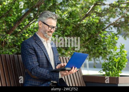 Elegante uomo d'affari senior con occhiali che utilizza un tablet digitale seduto su una panchina circondata dal verde. Foto Stock