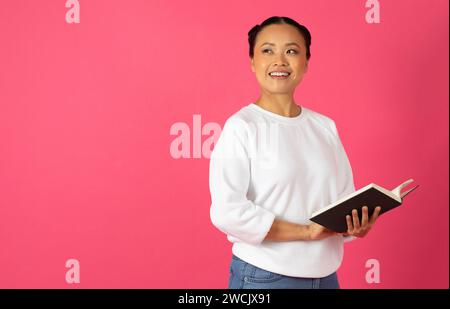 Felice giovane donna asiatica che tiene un libro aperto e sorride Foto Stock