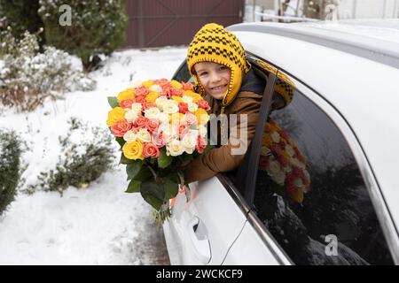 un ragazzo carino tiene in mano un grande mazzo di fiori appesi al finestrino di un'auto in inverno. Una piacevole sorpresa floreale per la mamma. Il figlio dà a sua madre un anticipo di compleanno Foto Stock