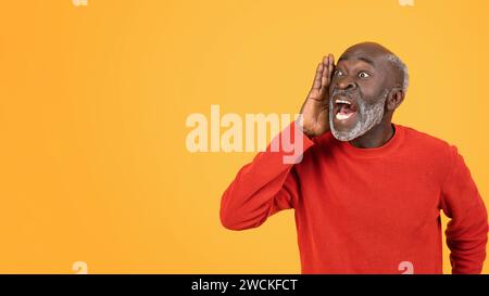 Un uomo anziano gioioso con un maglione rosso che condivide ad alta voce notizie o pettegolezzi emozionanti, mano a bocca Foto Stock