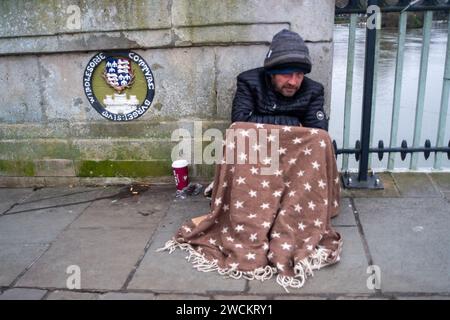 Eton, Windsor, Regno Unito. 16 gennaio 2024. Un senzatetto siede tristemente sul Windsor Bridge a Windsor, nel Berkshire, con una coperta che copre le ginocchia in una giornata fredda e gelida. L'uomo chiedeva ai passanti se potevano risparmiare qualche cambiamento. Il protocollo di emergenza tempo grave è attualmente in vigore nel Royal Borough di Windsor & Maidenhead il che significa che le persone senza tetto possono ottenere un alloggio temporaneo per la notte. Credito: Maureen McLean/Alamy Live News Foto Stock