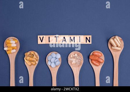 Vitamine diverse in cucchiai di legno con testo su sfondo grigio. Pillole, farmaci, integratori, medicina, farmacia e concetto sanitario. Foto Stock