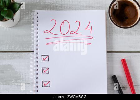 2024 obiettivi testo scritto a mano nel notebook con lista di controllo, tazza da caffè e piante succulente su legno. Vista della tabella superiore. Piano di Capodanno, risoluzioni, idee. Foto Stock