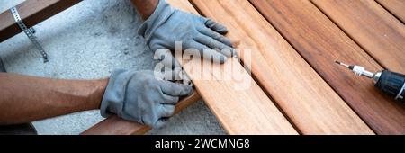 Mani maschili in guanti da lavoro protettivi posizionando un distanziatore in plastica tra tavole di legno mentre posa un nuovo patio esterno. Immagine grandangolare. Foto Stock