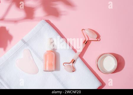 Rullo cosmetico per il viso, raschietto gua Sha, bottiglia di siero e vaso panna su un asciugamano bianco su sfondo rosa con ombre floreali. Concetto di centro benessere domestico. La via principale Foto Stock