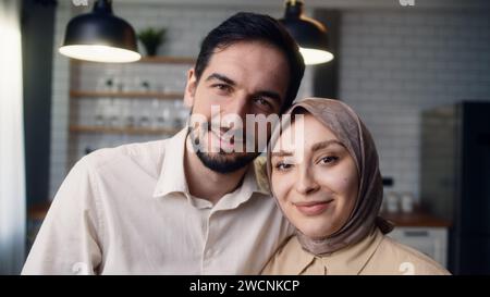 Ravvicinati la giovane coppia sposata che si abbracciano in cucina, sorridono e guardano la telecamera Foto Stock