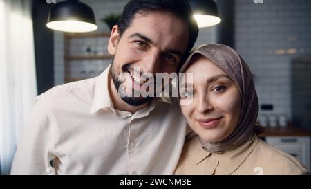 Ravvicinati la giovane coppia sposata che si abbracciano in cucina, sorridono e guardano la telecamera Foto Stock