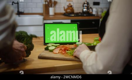 Computer tablet con schermo verde fittizio con display cromatico sul bancone della cucina, sposi che tritano verdure per insalata Foto Stock