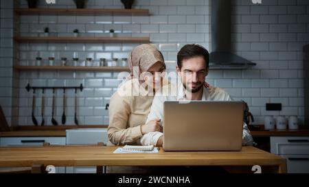 Un giovane uomo sposato sta lavorando al suo portatile in cucina quando sua moglie viene da dietro, lo abbraccia e sorride Foto Stock