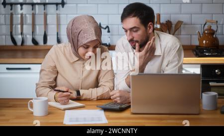 Giovani coppie sedute in cucina con il laptop pagano le bollette, utilizzano la calcolatrice per il conteggio delle spese, gestiscono le finanze, controllano il budget, si sentono stressati Foto Stock