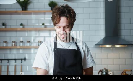 Ritratto di un uomo felice con capelli ricci con un grembiule a casa in una cucina moderna Foto Stock