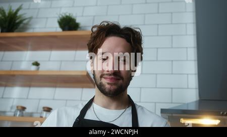 Ritratto con angolo di ripresa basso di un giovane uomo con i capelli ricci che guarda la fotocamera in cucina mentre si appoggia contro il bancone della cucina Foto Stock