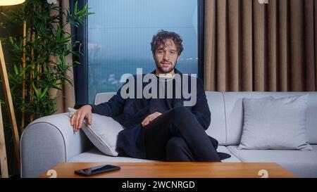 Bell'uomo d'affari dai capelli ricci che parla, guarda la macchina fotografica che effettua una chiamata in conferenza mentre si siede sul divano a casa. Concetto Videochat Foto Stock