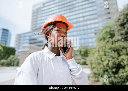 Donna nera seria con casco che parla allo smartphone di giorno Foto Stock