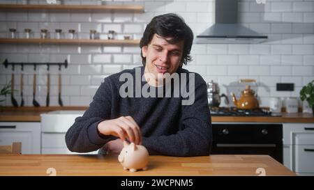 Un uomo felice seduto in una cucina moderna a casa mette un paio di monete nel salvadanaio Foto Stock