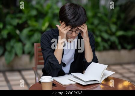 Un uomo asiatico stressato e sconvolto sta lavorando al suo incarico a un tavolo all'aperto, soffrendo di mal di testa, sentendosi troppo occupato. Foto Stock